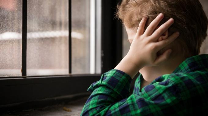Liten gutt som står bak vinduet i trist humør. Trist tenåring som ser i vinduet og lukker ørene med hendene. Ulykkelig barn i en rutet skjorte. Hjemme alene. Opprørt.